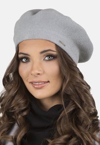 Berretto a maglia grigio con una texture morbida, forma rotonda e piccolo accento metallico di lato. Indossato con un top nero a collo alto.