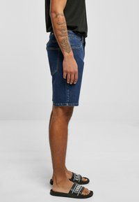 Shorts en denim bleu foncé, présentant une coupe classique, une jambe droite et des ourlets cousus. Portés avec des sandales noires affichant une lettrage blanc.
