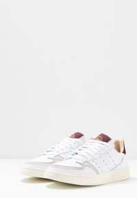 Vita sneakers med beige accenter, överdel av läder, perforerade detaljer och gummisula. Har en vinröd hälflik och snören.