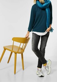 Person med teal färgad scarf och tröja, svarta jeans och vita sneakers står med ena handen på en senapsgul trästol.