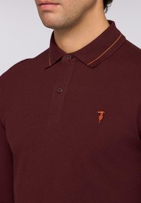 Polo di colore marrone con colletto corto, due bottoni, cuciture a contrasto arancioni e un piccolo logo ricamato sul petto, realizzato in tessuto testurizzato.