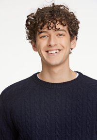 Dunkelblauer Strickpullover mit Rundhalsausschnitt. Verfügt über ein strukturiertes Muster und gerippte Bündchen, getragen über einem weißen Hemd. Das Modell lächelt.