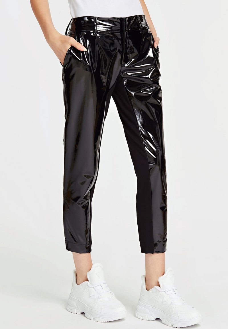 Persoon in zwarte glanzende vinylbroek met zakken en witte chunky sneakers, staand tegen een effen lichte achtergrond.