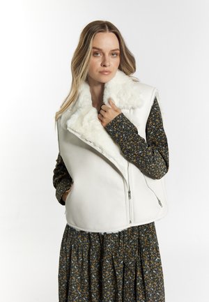 Witte mouwloze vest met faux fur kraag, voorzien van ritsluiting en contrasterende gedessineerde stof op de mouwen, tegen een eenvoudige achtergrond.