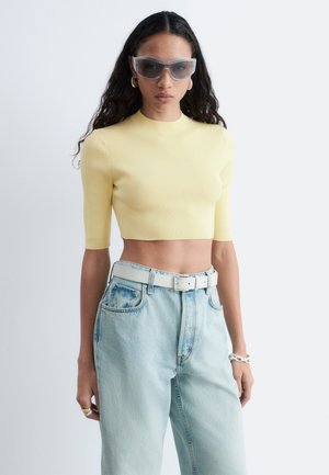 Kvinde med langt mørkt hår iført klare oversized solbriller, en gul cropped top, lyseblå jeans med et hvidt bælte og smykker.