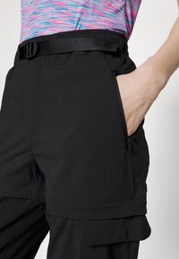 Personne portant un pantalon cargo noir zippé avec une ceinture noire et un haut coloré rose et bleu, main dans la poche droite.
