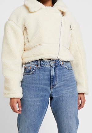 Personne portant une veste sherpa blanche courte avec des fermetures éclair argentées et un jean bleu taille haute sur un fond uni.