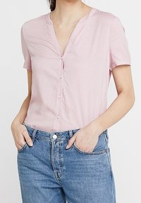 Blouse rose à manches courtes avec un col en V, fermeture à boutons et texture subtile. Portée avec un denim bleu clair, doté de poches et d'une coupe décontractée.