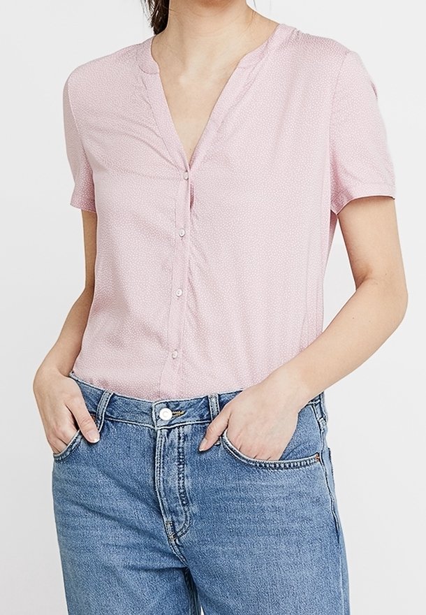 Blouse rose à manches courtes avec un col en V, fermeture à boutons et texture subtile. Portée avec un denim bleu clair, doté de poches et d'une coupe décontractée.
