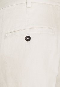 Tissu blanc texturé d'un pantalon avec une poche arrière ornée d'un seul bouton noir et un petit accent cousu au-dessus de la poche.