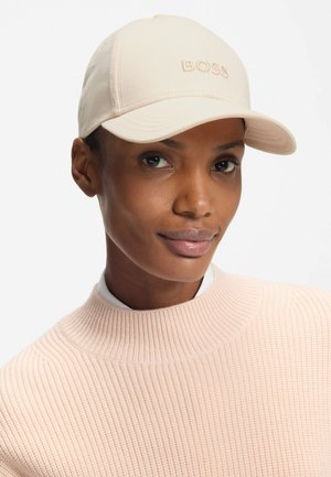 Beige cap with flat brim and raised embroidery of "BOSS" on front. Worn with a textured pink sweater. Smooth fabric, fitted design.