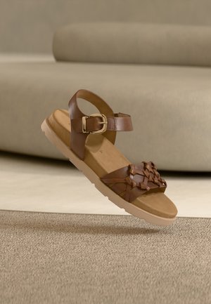 Brown leather sandal featuring a woven strap design, ankle buckle, and a light-colored sole. The footbed is cushioned for comfort.