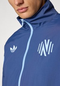 Marineblå nylonjakke med høy krage, lyseblå glidelås og logoer. Har et minimalistisk design, glatt tekstur og ingen synlige mønstre.