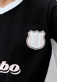 Černý dres s černým a světle modrým štítovým logem, který obsahuje nápisy "PALAAIS" a "1878", a bílými akcenty na límci.