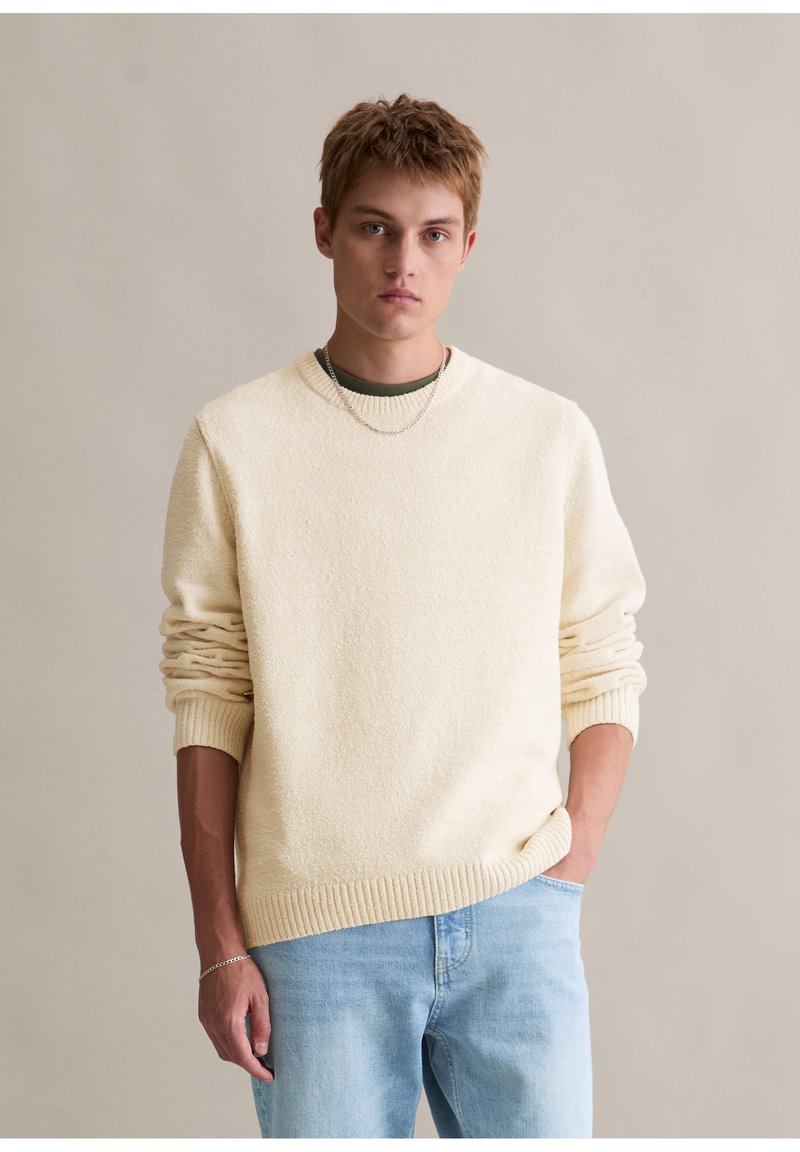 Cremetexturiertes Sweatshirt mit gerippten Bündchen und Saum, getragen über einem dunkelgrünen Hemd. Kombiniert mit hellblauen Jeans.