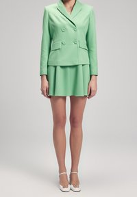 Žena v mátově zeleném dvouřadém saku a odpovídající mini sukni s bílými podpatky, stojící na jednobarevném šedém pozadí.