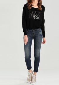 Schwarzer Sweatshirt mit Pailletten-Text "LOVE", kombiniert mit ausgewaschenen blauen Skinny-Jeans. Das Modell trägt beige Schnürsneaker. Sanfte Texturen.