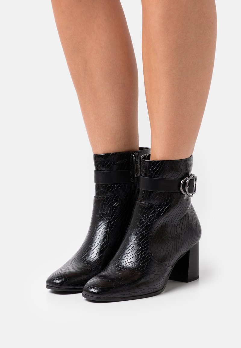 Bottes noires à la cheville en cuir texturé, talons carrés, boucles latérales et fermetures éclair, portées sur des jambes nues sur un fond clair uni.