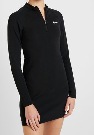 Kobieta ubrana w czarną, sportową sukienkę z długim rękawem i suwakiem, z białym logo Nike na piersi na jednolitym tle.