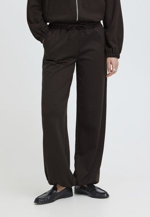 Osoba oblečená v čiernych nohaviciach s širokými nohavicami a sťahovacím pásom, čiernych mokasínach a čiernej bunde na zips, stojaca pred jednoduchým pozadím.