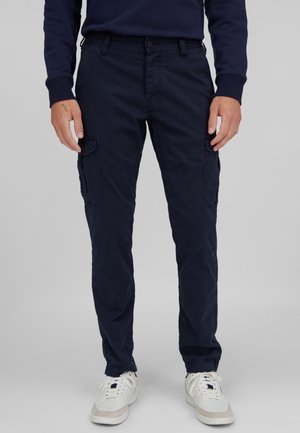 Hombre vestido con pantalones cargo azul marino y zapatillas blancas, de pie frente a un fondo gris liso.