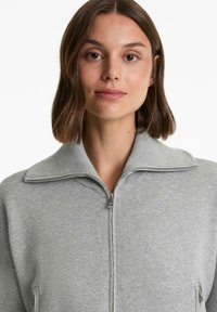 Vrouw met kort bruin haar, die een lichtgrijze ritstrui met een brede kraag draagt, tegen een effen witte achtergrond.