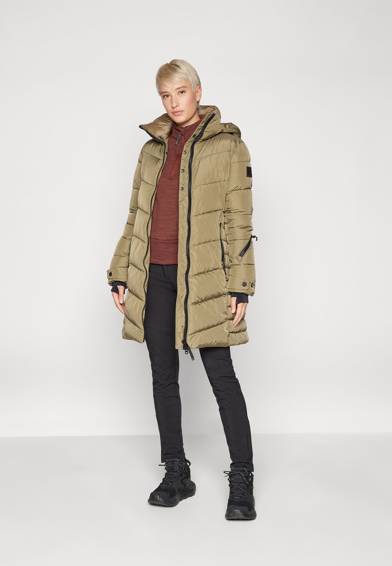 Leichte, olivgrüne Pufferjacke mit Kapuze, Reißverschluss und mehreren Taschen. Über einem bordeauxroten Oberteil und schwarzen Hosen getragen. Strukturierte Oberfläche.