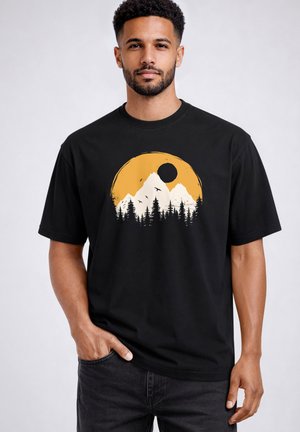 Man with short curly hair and beard wearing black T-shirt with orange sun, white mountains, pine trees, and flying birds graphic.