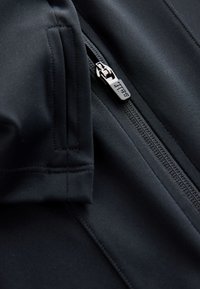 Zwarte jas met rits, gemaakt van een gladde, rekbare stof, met een close-up van de rits en dubbel gestikte naden voor duurzaamheid.