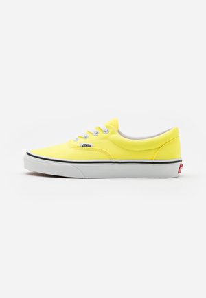 Baskets en toile jaunes avec lacets et semelle blancs. Présente une bande noire le long du bord et un petit logo "Vans" sur le côté. Design classique à basse hauteur.