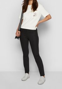 Witte polo met korte mouwen, voorzien van een borstpatch, gecombineerd met zwarte slim-fit broek en witte sneakers. De outfit heeft een casual ontwerp.
