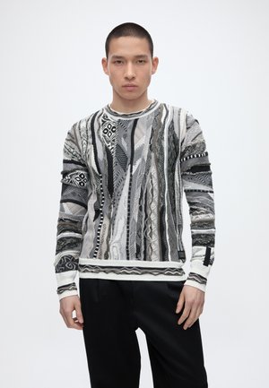 Jeune homme aux cheveux courts portant un pull en laine à motifs noirs, blancs et gris et un pantalon noir, debout devant un fond uni.