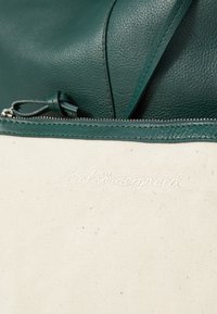 Zelena teksturirana kožna torba s patentnim zatvaračem, koja sadrži mekani bež platneni džep i izvezeni brand logo u bijeloj boji.