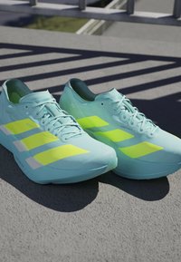 Light blue athletic shoes with yellow accents. Features a smooth finish, distinctive three-stripe design, and a cushioned sole.