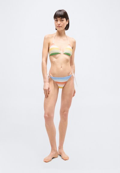 Γυναίκα που στέκεται ξυπόλυτη φορώντας μπικίνι με πολύχρωμο ζιγκ-ζαγκ μοτίβο, τοπ με χαλτέρ και σλιπ με δέσιμο στο πλάι, σε απλό ανοιχτόχρωμο φόντο.