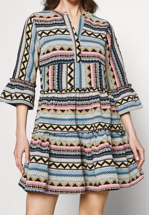 Mujer con un vestido colorido de patrones con diseños en zigzag y geométricos, mangas con volantes hasta el codo y falda con volantes en capas.