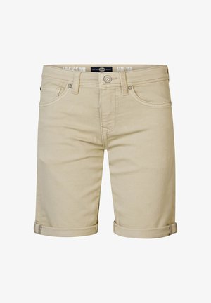 Bambino in piedi con una mano in tasca, indossa sneakers bianche, pantaloncini beige e una maglietta bianca con una toppa rotonda sul petto.