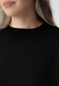 Sweatshirt en coton noir avec un col rond texturé, présentant un petit logo en relief à l'avant. Tissu lisse avec une coupe décontractée.