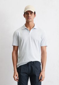 Lichtblauw polo-shirt met korte mouwen en een kraag, gecombineerd met donkere marineblauwe broek en een beige pet, tegen een gestructureerde witte achtergrond.