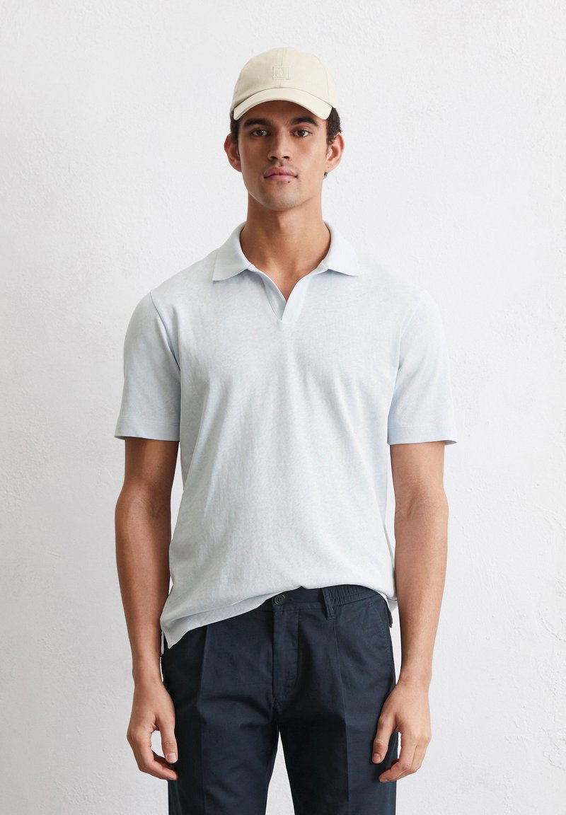 Lichtblauw polo-shirt met korte mouwen en een kraag, gecombineerd met donkere marineblauwe broek en een beige pet, tegen een gestructureerde witte achtergrond.