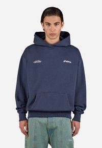 Young man wearing navy blue hoodie with small white text logos, paired with loose light blue jeans, standing against plain white background.
