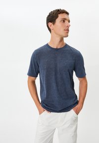 Mörkblå t-shirt med kort ärm och rundad halsringning, med en texturerad yta, bärs tillsammans med vita byxor, med händerna i fickorna, stående mot en enfärgad bakgrund.