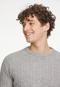 Szary sweter w splot kratek z okrągłym dekoltem, z teksturowanymi wzorami i miękkim wyglądem, noszony przez uśmiechającą się osobę z kręconymi włosami.