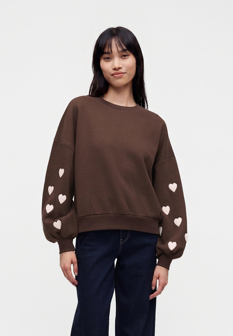 Jeune femme porte un sweat-shirt marron avec des motifs de cœurs roses sur les manches bouffantes et un jean bleu foncé, debout devant un fond uni.