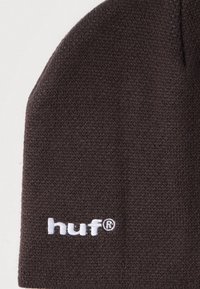 Sötétbarna kötött sapka, alján fehér hímzett "huf®" logóval, világos háttéren.
