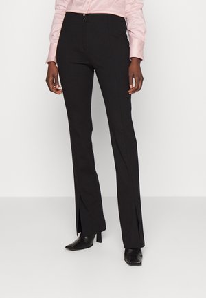 Osoba nosząca czarne spodnie garniturowe z wysokim stanem i przednimi rozcięciami, połączone z czarnymi szpiczastymi szpilkami i jasnoróżową koszulą zapinaną na guziki.