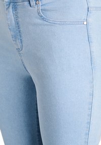 Svetlo modré džínsy z denimu majú priliehavý strih so šitými švami a funkčnými vreckami, predvádzajúc hladkú textúru a jemné blednutie.