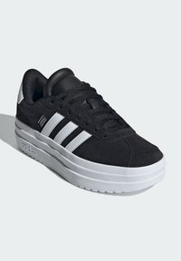 Sneaker en suede noir avec semelle en caoutchouc blanc, présentant trois rayures blanches, bout rond et languette rembourrée pour le confort.