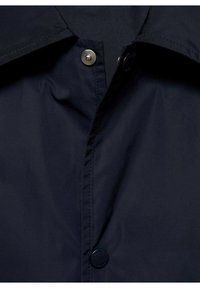 Veste légère bleu marine avec un col à rabat, une fermeture à pression et une texture lisse. Le tissu semble légèrement réfléchissant et résistant à l'eau.