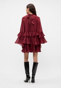 Burgunderfarget rufsete kjole med knyting i halsen, lagdelt stoff, gjennomsiktig tekstur, kombinert med høye svarte glansede støvler.